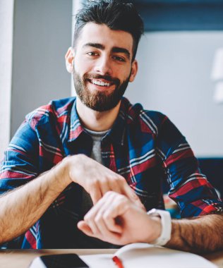 Üniversitede masada oturan akıllı saatin sergilenmesi sırasında kameraya gülümseyen pozitif sakallı genç adamın portresi. Rahat giyim giymiş neşeli hipster adam kol saati üzerinde zaman yönetmek