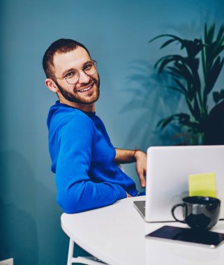 Ofisteki iş akışı sırasında kameraya bakan erkek çalışanın kesilip biçilmiş görüntüsünden memnun olan çok ırklı meslektaşlarıyla birlikte gözlüklü neşeli beyaz öğrencinin portresi.