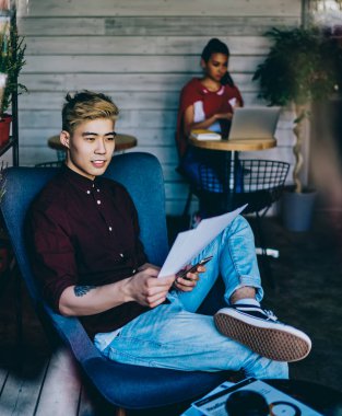 Düşünceli Japon hippi şık kıyafetler giyip kafeteryada rahat bir şezlongda dinleniyor. İçi rahat ve iş projesi üzerine kafa yoruyor ve kağıt üzerinde bilgi topluyor.