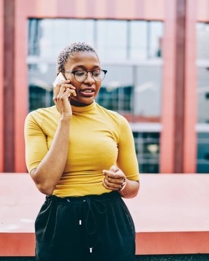 Gözlüklü Afrikalı Amerikalı kadın modern akıllı telefondaki telefon görüşmeleri sırasında şehir ortamında dolaşım bağlantısını kullanarak bazı bilgileri açıklıyor. Koyu tenli kadın cep telefonundan arıyor.