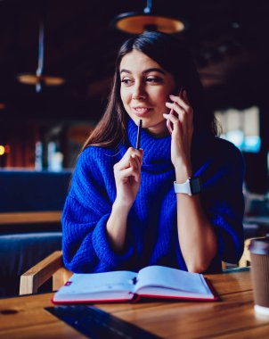 Kafeteryada dinlenirken modern akıllı telefon aracılığıyla arkadaşıyla olumlu sohbet eden neşeli hipster kız, kapalı mekanlarda 4g internet ile telefonla uygulama kullanarak arayan başarılı bir kadın