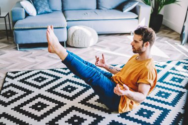 Sportif deneyimli genç adam, iç mekanda meditasyon sırasında halıya otururken bacağını kaldırıyor. Hippi adam sağlıklı bir hayat sürüyor ve modern apartman dairesinde sabah egzersizleri yapıyor.