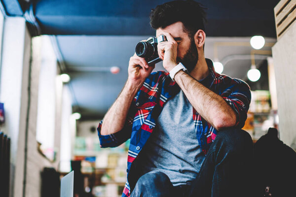 Bearded young man working photographer in studio and making photos on vintage camera.Hipster blogger holding modern camera and focusing to take picture sitting in coworking space