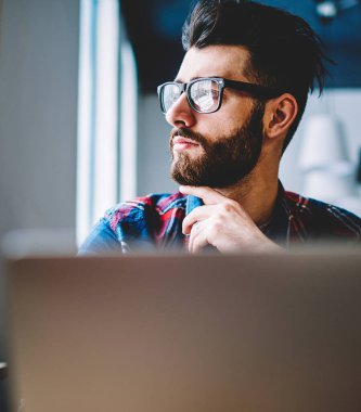 Modern dizüstü bilgisayarda serbest çalışırken uzak bakarken vizyon düzeltme için şık gözlük düşünceli sakallı genç adam. Hipster öğrencisinin yayınlanma kıfzıtları üzerine düşünmesi