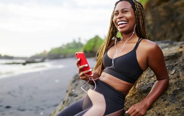 Deniz kıyısında dinlenen akıllı telefonbağlı kulaklıklar serin ses şarkıları zevk dreads ile mutlu afro amerikan koşucu, müzik dinleme eğitimi için aktif giysiler içinde pozitif koyu tenli jogger