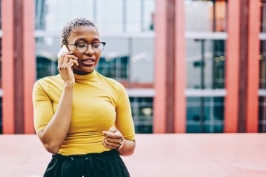 Gözlüklü Afrikalı Amerikalı kadın modern akıllı telefondaki telefon görüşmeleri sırasında şehir ortamında dolaşım bağlantısını kullanarak bazı bilgileri açıklıyor. Koyu tenli kadın cep telefonundan arıyor.