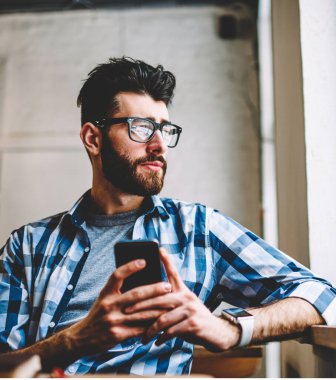 Akıllı telefon cihazındaki sosyal ağlardaki profili güncellerken gözlüklü hipster öğrencisinin pencereden dışarı bakmasından. Düşünceli sakallı genç adam cep telefonu online sohbet