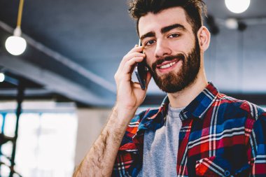Üniversite kütüphanesinde duran kameraya gülümserken modern akıllı telefonda konuşan yakışıklı neşeli genç adamın portresi. Pozitif hipster öğrenci edebiyat kitapları tutan telefonda arama