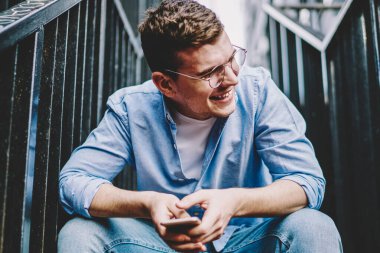 Sıradan giyinmiş, gülen, elinde akıllı telefon cihazıyla gezen pozitif bir genç. Cep telefonlu neşeli bir internet blogcusu merdivenlerde oturuyor.