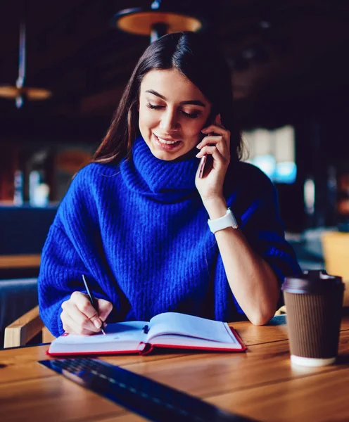 Cheerful woman with cellular phone calling to friend while studying at campus cafe, smiling hipster girl connected to 4g internet talking by telephone during writing notes and sitting at cafe