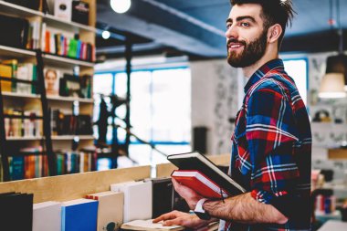 Pozitif yakışıklı genç adam boş zamanlarında okumak için edebiyat seçerken elinde kitap tutan şık gömlek giymiş. Akıllı sakallı hipster öğrenci kütüphanede ayakta uzakta bakıyor