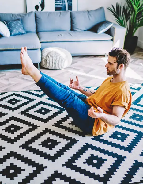 Experienced young man in sportive wear holding legs up sitting on carpet during meditation at home interior.Hipster guy lead healthy lifestyle and doing morning exercises in modern apartment