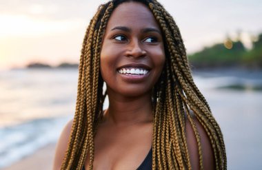 Sahilde sabah eğitimi öncesi dreads ile Neşeli afro amerikan koşucu, pozitif koyu tenli kadın aktif giyim sahil şeridinde ayakta giyinmiş ve yüzünde sevimli bir gülümseme ile uzağa bakıyor