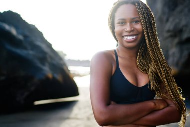 Neşeli afro amerikan genç kadın spor sevgilisi portre kamera önünde elleri çapraz iken gülümseyen, mutlu kadın arka planda kayalar ile açık havada ayakta egzersiz için aktif üst giymiş