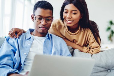 Gülümseyen Afro-Amerikan kadın, oturma odasında dizüstü bilgisayarla oturan, web mağazasında birlikte alışveriş yapan, siyah tenli, pozitif bir çift, serbest çalışan kocasına bakıyor.