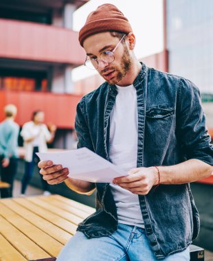 Düşünceli genç adam gündelik giysiler giymiş, elinde kağıt dersleri tutuyor ve metin bilgileri okuyor şehir ortamında seminer için hazırlanıyor. Hipster öğrenci raporlarını dışarıda kontrol ediyor.