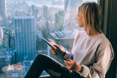 Sıradan giyinen şık sarışın kadın şehir merkezindeki Empire State binasının manzarasını izlerken pencere eşiğinde oturuyordu. New York 'un popüler eğlenceleri. Dijital Göçebe