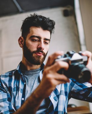 Penisli profesyonel fotoğrafçı vintage cihaz tutan ve kendi stüdyosunda oturan fotoğrafları izliyor. Sakallı hipster adam rahat giyim giymiş kamera resimleri görüntüleme ve ayar yapma