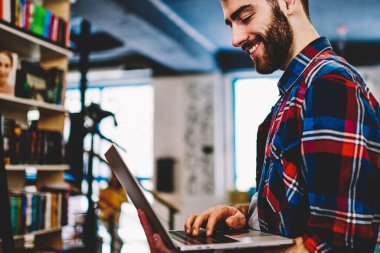 Elinde dizüstü bilgisayar tutan sakallı neşeli genç adam ve modern kütüphanede kablosuz internet bağlantısı kullanarak uygulama yükleyerek. Netbook online sohbet Pozitif hipster adam