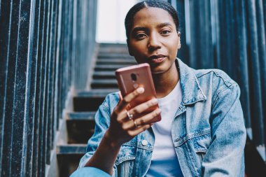 Güzel Afro-Amerikan genç bir kadının portresi kameraya bakıyor, elinde akıllı telefon cihazı var ve 4G internet bağlantısı üzerinden uygulama yüklüyor. Yakışıklı, koyu tenli hippi kız cep telefonuyla sohbet ediyor.