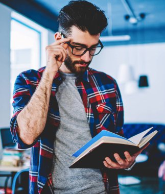 Görme düzeltme için gözlük pensive akıllı hipster öğrenci elinde ders kitabı tutan ve ders yazmak için bilgi okuma. Kütüphanede ayakta rahat giyim konsantre genç adam