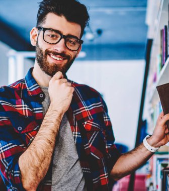 Üniversitede kitaplıktan edebiyat kitabı seçerken kameraya gülümseyen gözlüklü başarılı sakallı hipster öğrencisinin portresi. Kütüphanede duran rahat gömlek pozitif genç adam