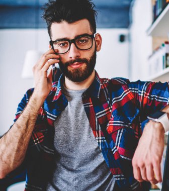 Hipster öğrencisinin gözlüklü portresi, kameraya bakarken kütüphanedeki kitaplığın yanındaki merdivene el ile eğildi. Sakallı mae kütüphaneci üniversitede ayakta modern akıllı telefon çağrı