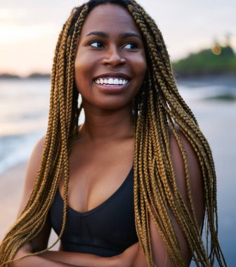 Sahilde sabah eğitimi öncesi dreads ile Neşeli afro amerikan koşucu, pozitif koyu tenli kadın aktif giyim sahil şeridinde ayakta giyinmiş ve yüzünde sevimli bir gülümseme ile uzağa bakıyor