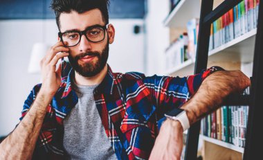 Hipster öğrencisinin gözlüklü portresi, kameraya bakarken kütüphanedeki kitaplığın yanındaki merdivene el ile eğildi. Sakallı mae kütüphaneci üniversitede ayakta modern akıllı telefon çağrı