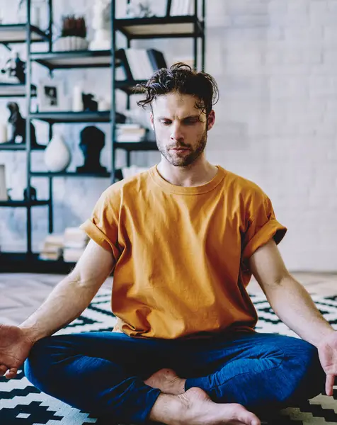 Concentrated young bearded man in active wear sitting in lotus pose during morning training on comfortable carpet in modern apartment.Calm hipster guy engaged in yoga at home interior