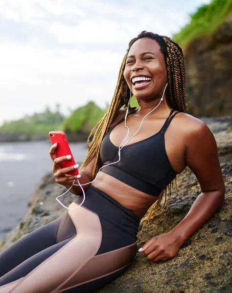 Happy afro american runner with dreads enjoying cool audio songs in earphones connected to smartphone resting on sea coast, positive dark skinned jogger in active clothes for training listening music