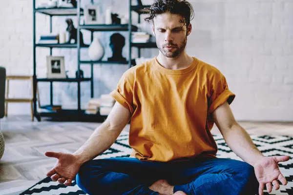 Aktif görevde olan genç sakallı bir adam sabah nilüferde poz verirken modern apartmandaki rahat halı üzerinde antrenman yapıyor. Evde yoga yapan sakin bir hippi çocuk.