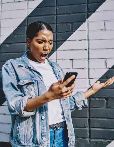 Angry african american young woman shocked with received notification with unhappy news standing in urban setting.Shocked dark skinned hipster girl has problem in mobile app on modern cellular