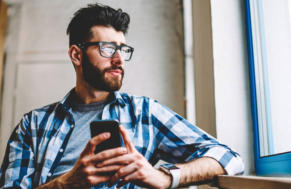 Pondering hipster student in eyeglasses looking out window while updating profile in social networks on smartphone device.Thoughtful bearded young man chatting online on mobile phone