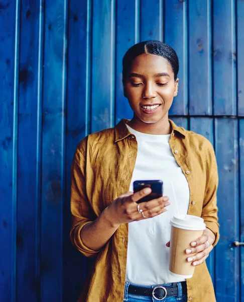 Cheerful african american young woman holding coffee to go and chatting online on mobile phone using public 4G internet standing against wall in urban setting.Positive dark skinned hipster blogger