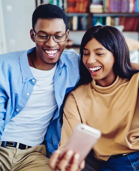 stock image Cheerful dark skinned hipster couple making funny faces posing for selfie and using smartphone app, happy african american girl and guy sitting on sofa spending time at home making picture together