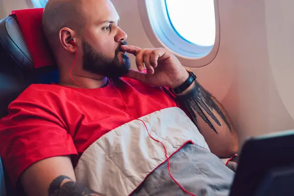 Thoughtful caucasian male passenger looking in large flight window sitting in comfortable first business class seat beginning vacations. Young man tourist listen music in earphones with entertainment