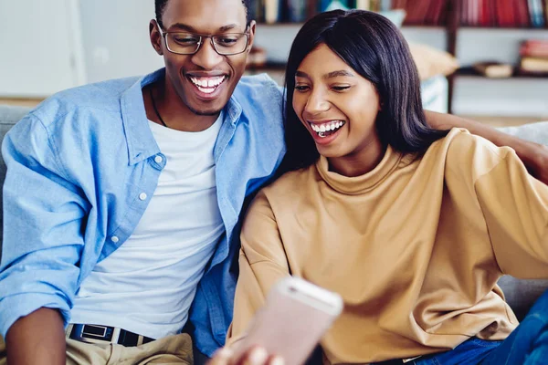 Neşeli koyu tenli hippi çift selfie için komik suratlar yapıyor ve akıllı telefon uygulaması kullanıyorlar. Mutlu Afro-Amerikan kız ve erkek kanepede oturup birlikte fotoğraf çekiyorlar.