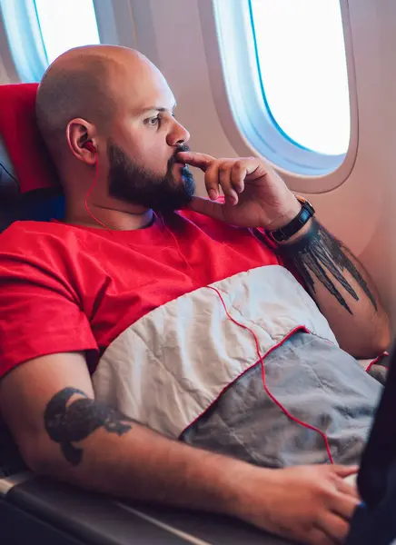Thoughtful caucasian male passenger looking in large flight window sitting in comfortable first business class seat beginning vacations. Young man tourist listen music in earphones with entertainment