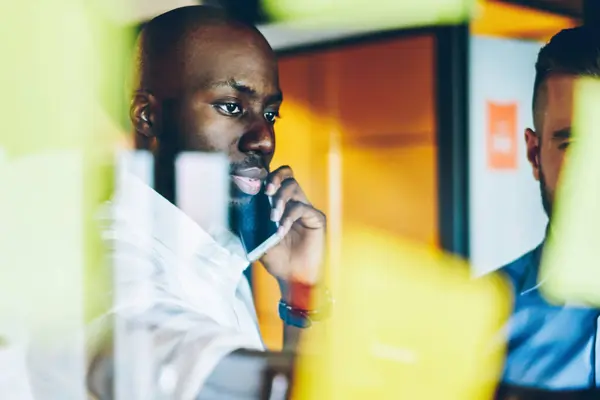 Pensive dark skinned proud ceo in formal wear calling via phone call on modern smartphone while standing with colleagues behind glass office wall with colorful stickers. Meeting of workers in office