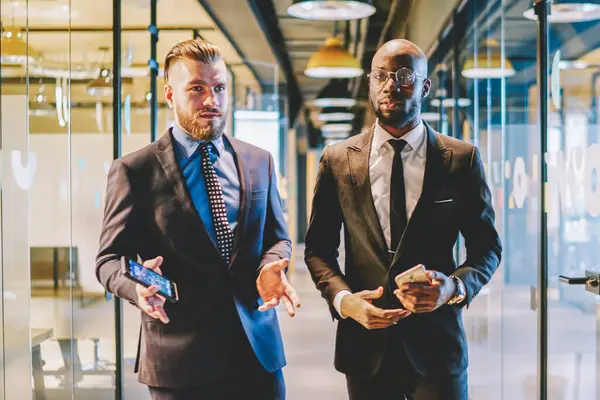 Caucasian experienced proud ceo with dark skinned business partner dressed in suit going on office corridor and communicating with each other about development of project and successful cooperation