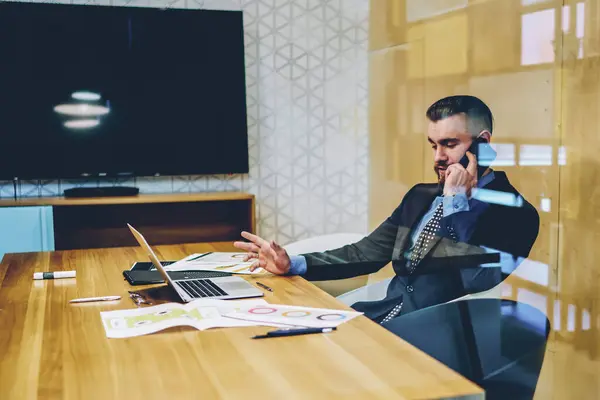Confident owner of financial company working at netbook in own office and calling on smartphone to discuss business plan of new project. Male entrepreneur in suit talking on digital smartphone