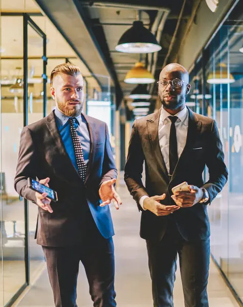 Caucasian experienced proud ceo with dark skinned business partner dressed in suit going on office corridor and communicating with each other about development of project and successful cooperation