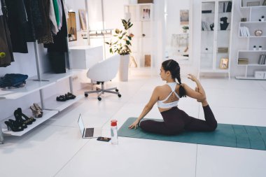 Oturma odasında güvercin yoga pozuna hazırlanan dizüstü bilgisayara bakarken el ve ayak üzerinde mindere oturmuş genç bir kadının yan görüntüsü.