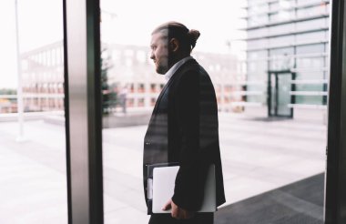 Modern binanın terasında elinde dizüstü bilgisayarla gölgede duran ve gün ışığında şehir merkezindeki bulanık manzarayı izleyen sakallı erkek girişimcinin camdan manzarası.