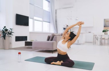 Padmasana 'da su şişesi ile minderde oturan bir kadının yan görüntüsü. Kapalı gözlerle poz veriyor ve kollarını başlarının üstüne uzatarak el ele tutuşuyor ve oturma odasında yoga yapıyor.