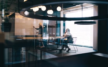 Masada oturan kadın ve toplantı odasında çalışan erkek ve cam kapılar, odanın karşısındaki araba sokağını ve perdeyi yansıtıyor.