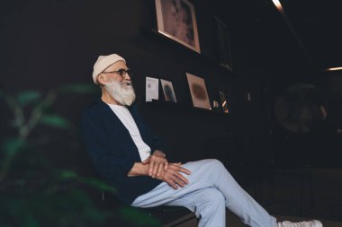 Şapkalı yaşlı bir adamın karanlık odada bacak bacak üstüne atarak oturması ve tek başına zaman geçirirken başka tarafa bakması.