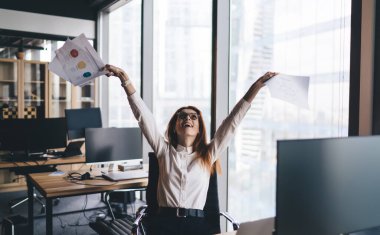Modern ofiste bilgisayarlı pencerenin yanında otururken resmi giysiler içindeki heyecanlı kadın ofis çalışanı belgelerle kollarını kaldırıyor.