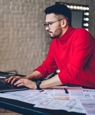 Gözlüklü ciddi bir genç, evdeki uzaktan çalışma sırasında netbook üzerinden araştırma yapan, kablosuz internet kullanan, konsantre hipster adam, modern dizüstü bilgisayardan bilgileri kontrol ediyor.